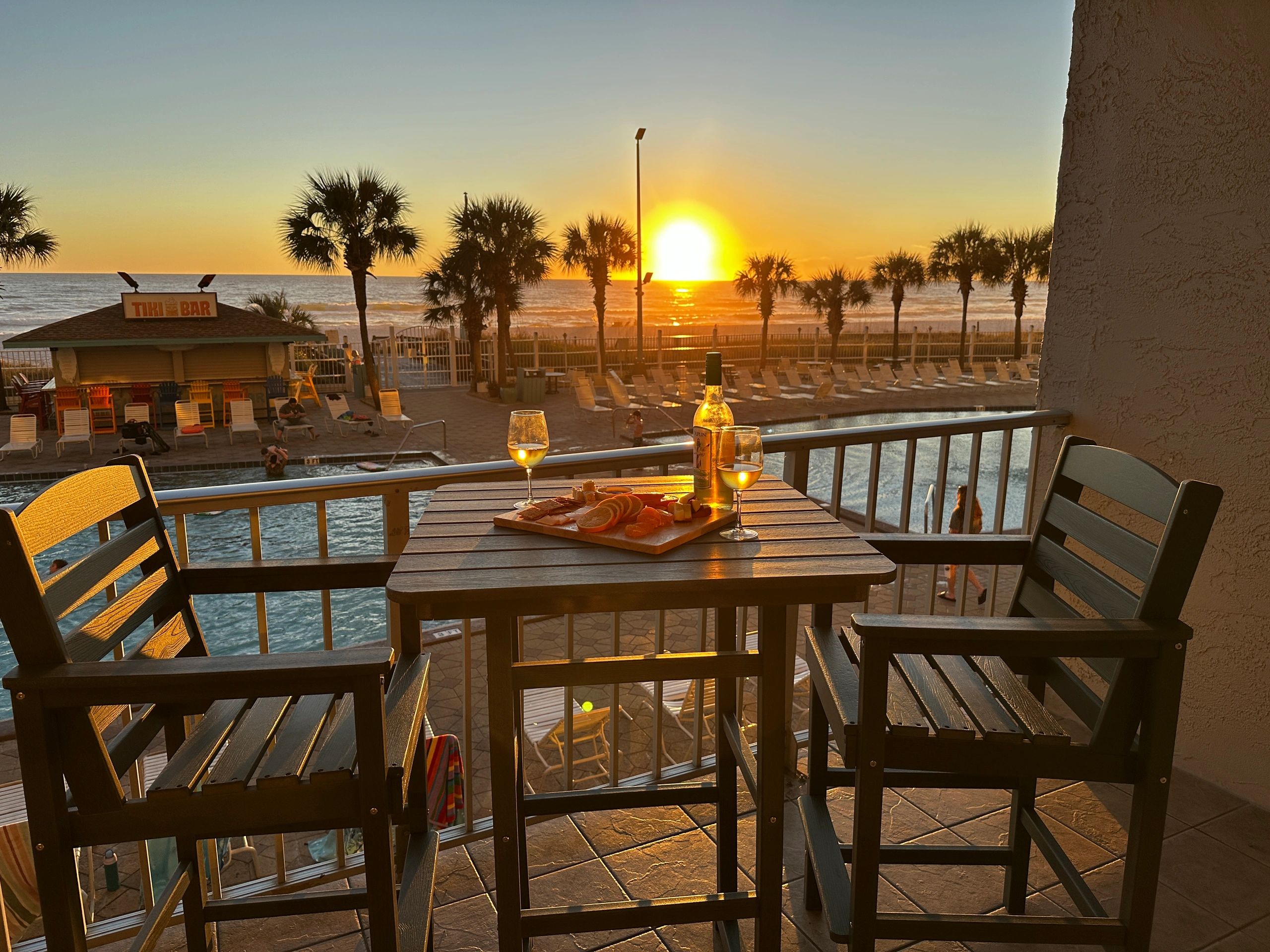 Sunset view from a balcony with wine and snacks overlooking a pool and beach.