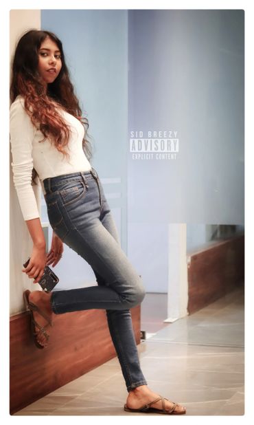 Young woman leaning against a wall holding a phone, dressed casually in jeans and a white top.