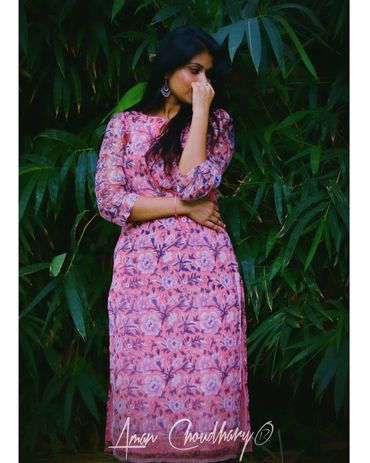 Woman in a pink floral dress standing thoughtfully among green foliage.