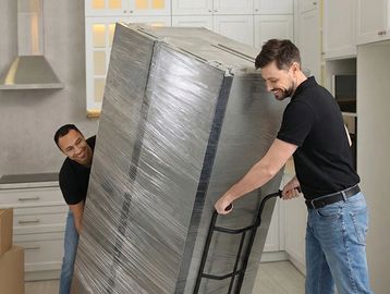 Two men moving a large wrapped appliance on a dolly inside a kitchen.