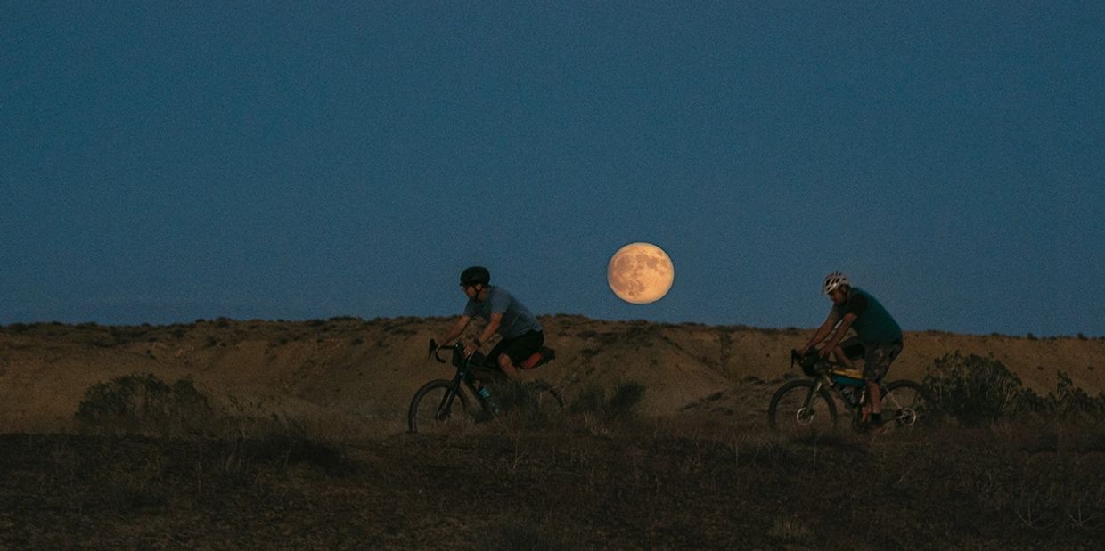 Two cyclists ride under a full moon in a desert landscape at dusk.
