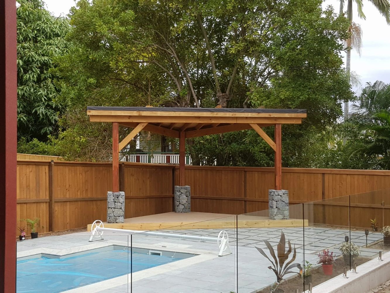 A wooden pergola near a pool with stone pillars and glass fencing.