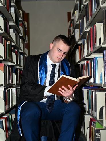 Graduate in academic gown reading a book in a library aisle.