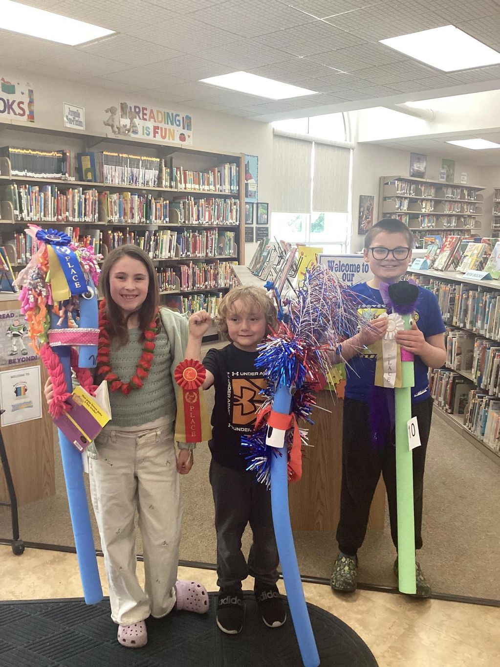 Three children proudly display decorated pool noodles and ribbons in a library.