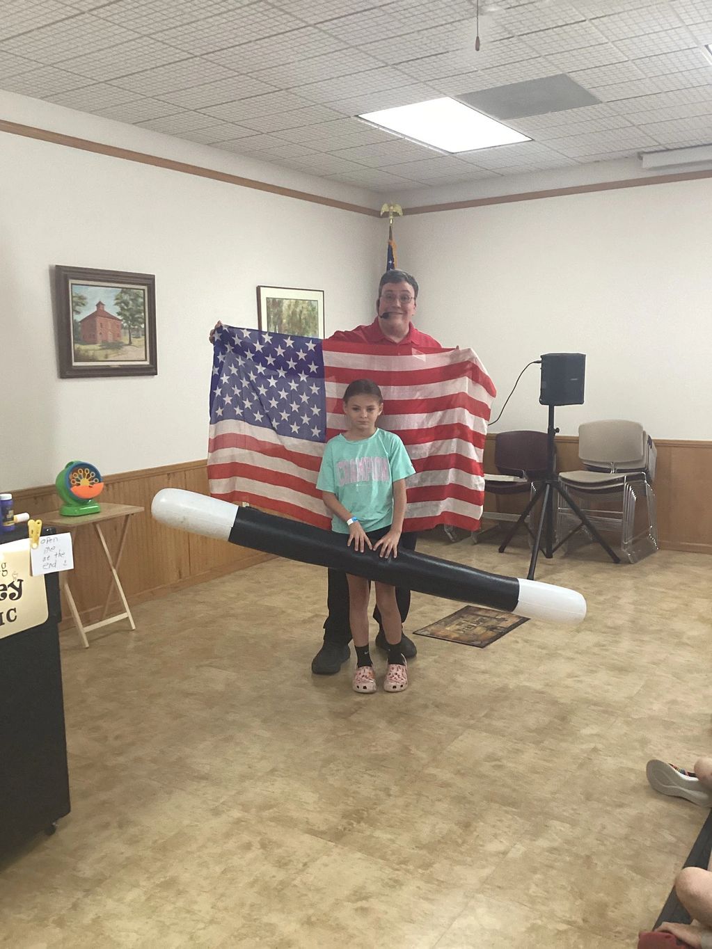 A man and a girl pose with a giant magic wand and an American flag indoors.