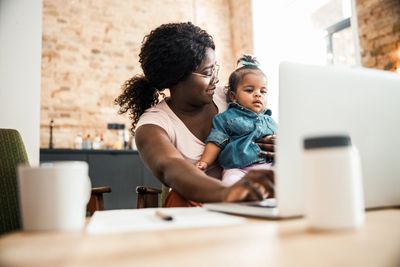 Woman holding baby, typing on laptop.