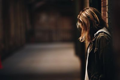 Young woman leaning against wall in a long, darkened street, with her head down, hair hiding face.
