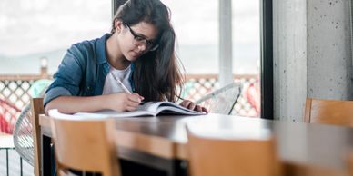 Young woman with glasses concentrating, leaning over notebook with pen in hand.