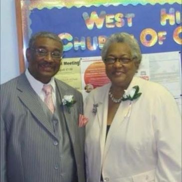 Elderly couple dressed formally, smiling at the camera in front of a church bulletin board.