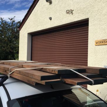 Wooden planks secured on a car roof rack outside a workshop.