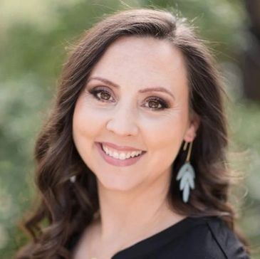Smiling woman with long wavy hair and earrings in an outdoor setting.