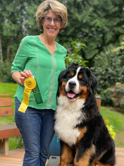 A woman hold her Rally Obedience ribbons as she stands next to her Bernese Mountain Dog
