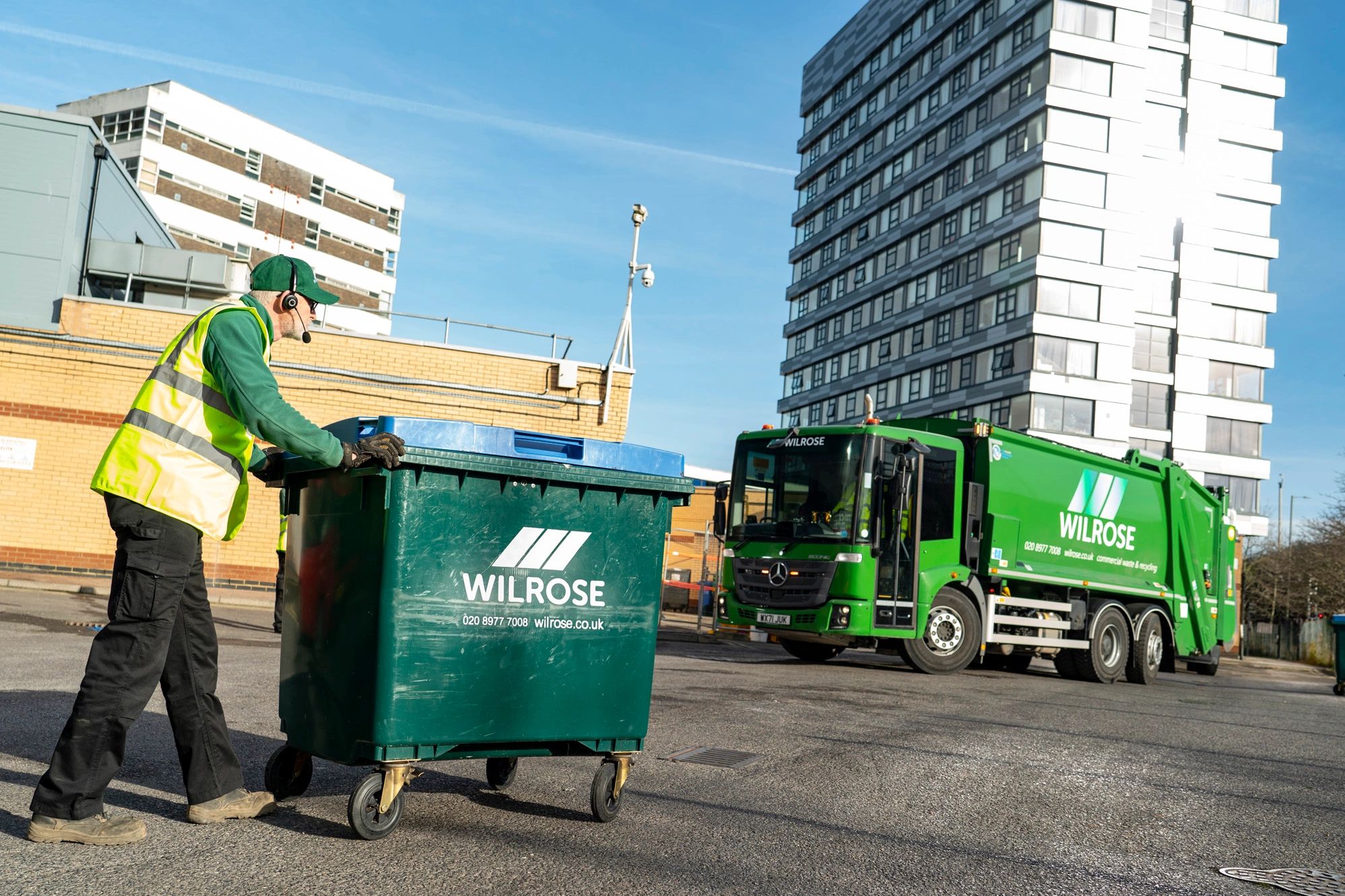 Driver pushing 1100L commercial waste wheelie bin towards green dustcart.