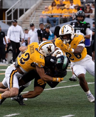 Two yellow uniform defenders tackle a black uniform football player.