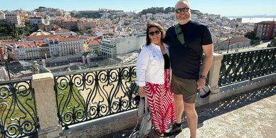 Couple posing on a scenic balcony overlooking a sunlit cityscape with historic buildings.