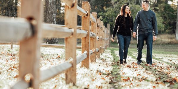 Couple walking hand-in-hand along a wooden fence in a sunlit park.
