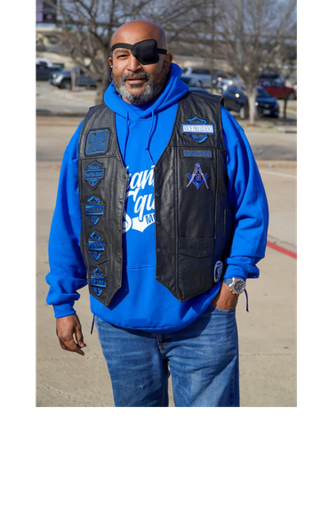Man wearing a blue hoodie, leather vest with patches, and an eye patch outdoors.