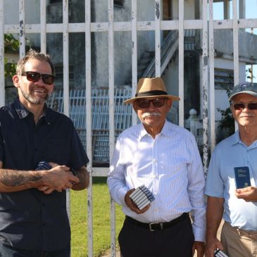 Three men smiling and holding items outdoors near a fence.