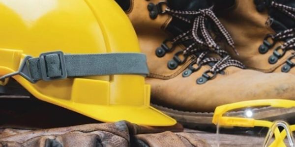 Safety gear including a yellow hard hat, gloves, boots, and protective glasses on wooden surface.