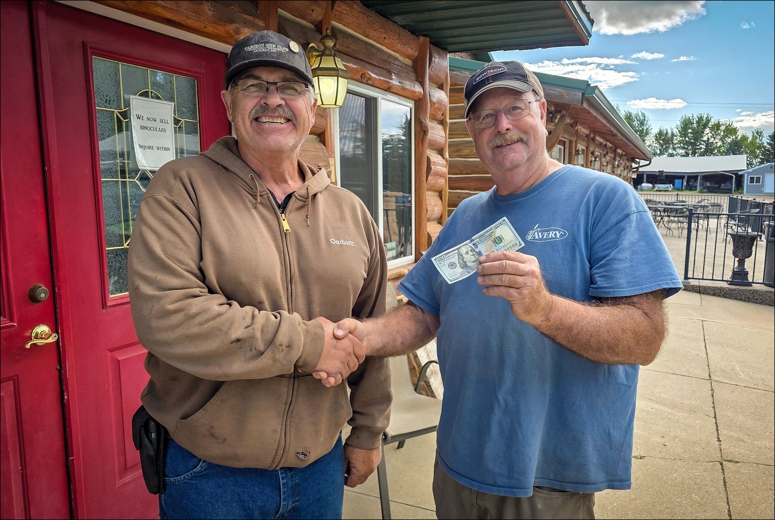 Two men smiling and shaking hands outside a rustic building, one holding cash.