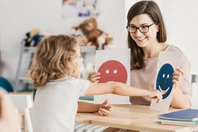 Lady and child interacting. Lady holding up smiley face pictures and child pointing to smiley face.