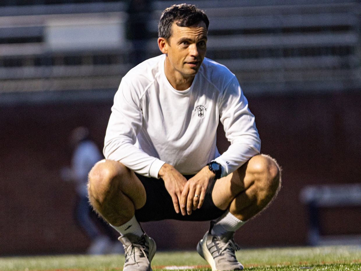 A man squatting on a sports field wearing a white long-sleeve shirt and gray sneakers.