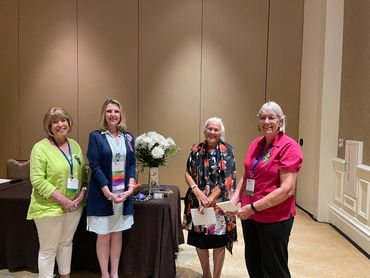 Four women standing indoors near a table with flowers.