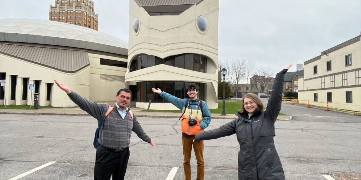 Three people posing happily in front of a uniquely designed building on a cloudy day.