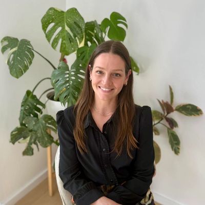 Woman with long brown hair sitting and smiling indoors with green plants in the background.