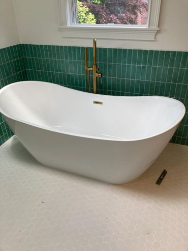 Modern white freestanding bathtub with green tiled wall and gold faucet.