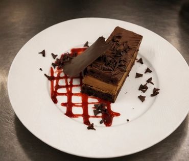 A slice of chocolate mousse cake with chocolate shavings and raspberry sauce.