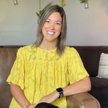 Smiling woman in a bright yellow blouse sitting on a brown chair.