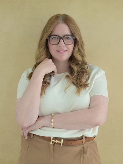 Femme souriante aux lunettes, cheveux ondulés, portant un haut crème et un pantalon beige.