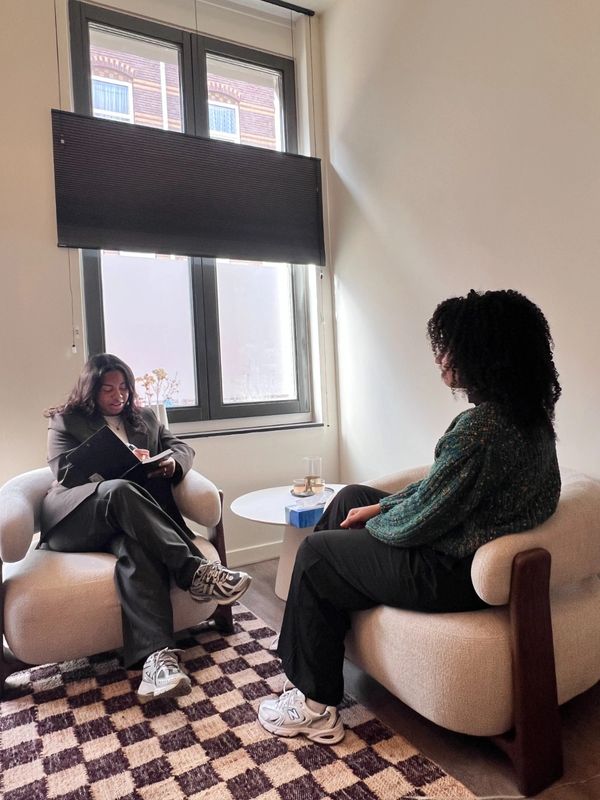 Two women sit in a cozy room engaged in a conversation, one taking notes.