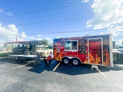 Chef Todd sitting in front of smoker trailer and All Fired Up BBQ Service trailer