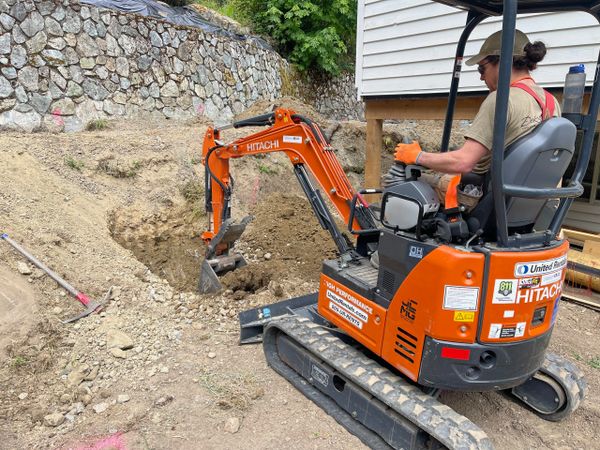 Person operating a mini excavator to dig a trench near a house.