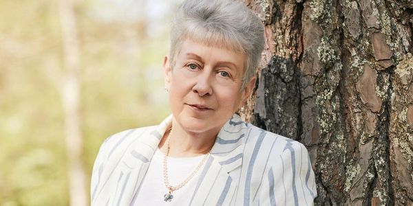 Older woman with gray hair, wearing a striped blazer, leaning against a tree.