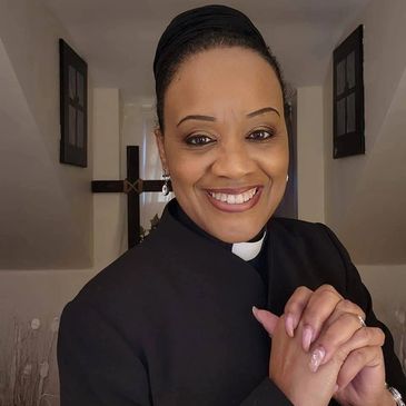 Smiling woman in clerical attire with a cross in the background.