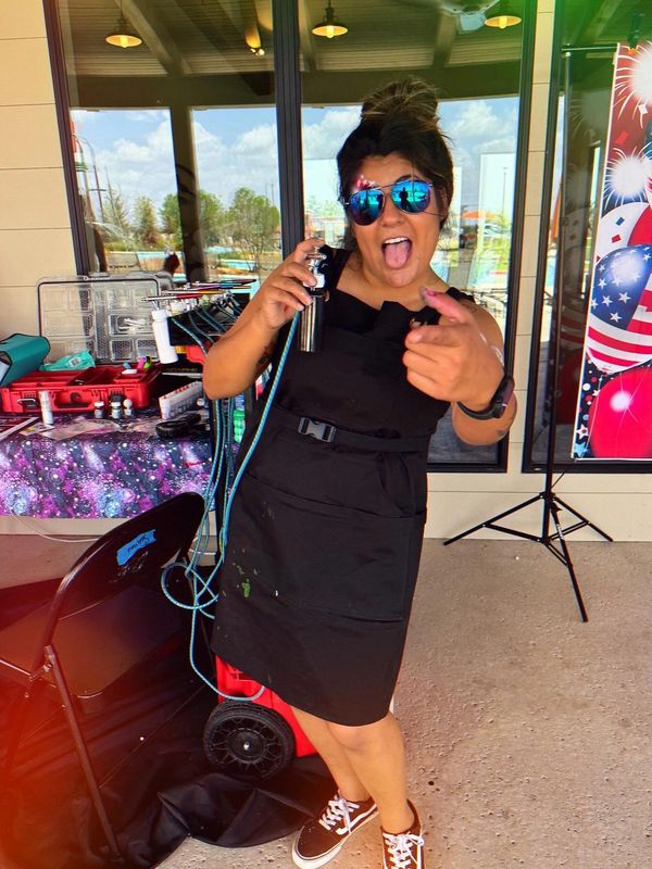 Woman in black apron playfully posing with airbrush equipment.
