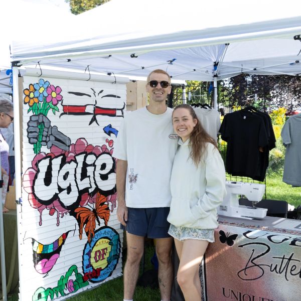 Two people smiling at a colorful outdoor booth with art and clothing.