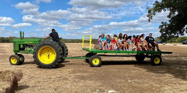 Children and adults riding a hay wagon pulled by a John Deere tractor on a sunny day.