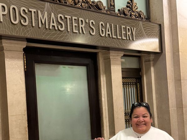 A smiling woman stands under the Postmaster's Gallery sign inside a building.