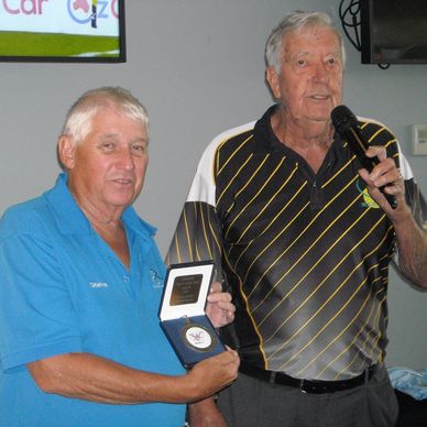 Two elderly men at an award ceremony, one holding an award and the other speaking into a microphone.