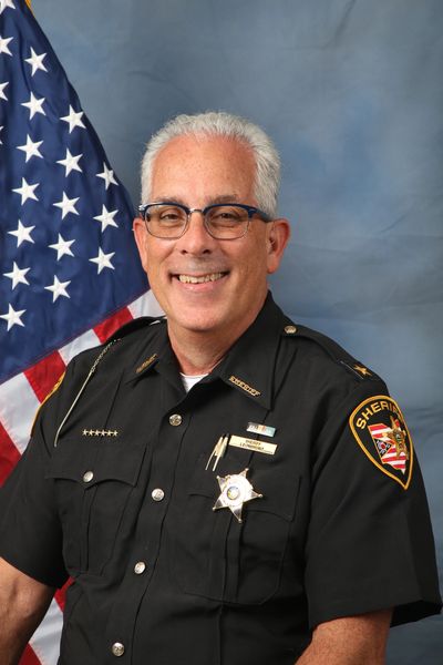 Smiling sheriff in uniform poses with American flag backdrop.
