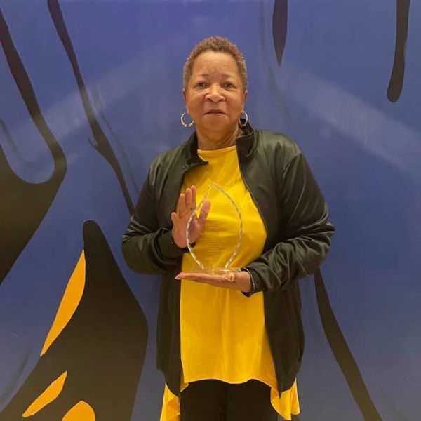 A woman in a yellow dress and black jacket holds a glass award.