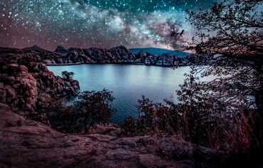 Starry night over a serene lake surrounded by rocky hills and trees.