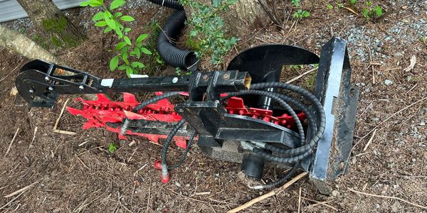 A red and black mechanical soil tiller lying on the ground near trees.