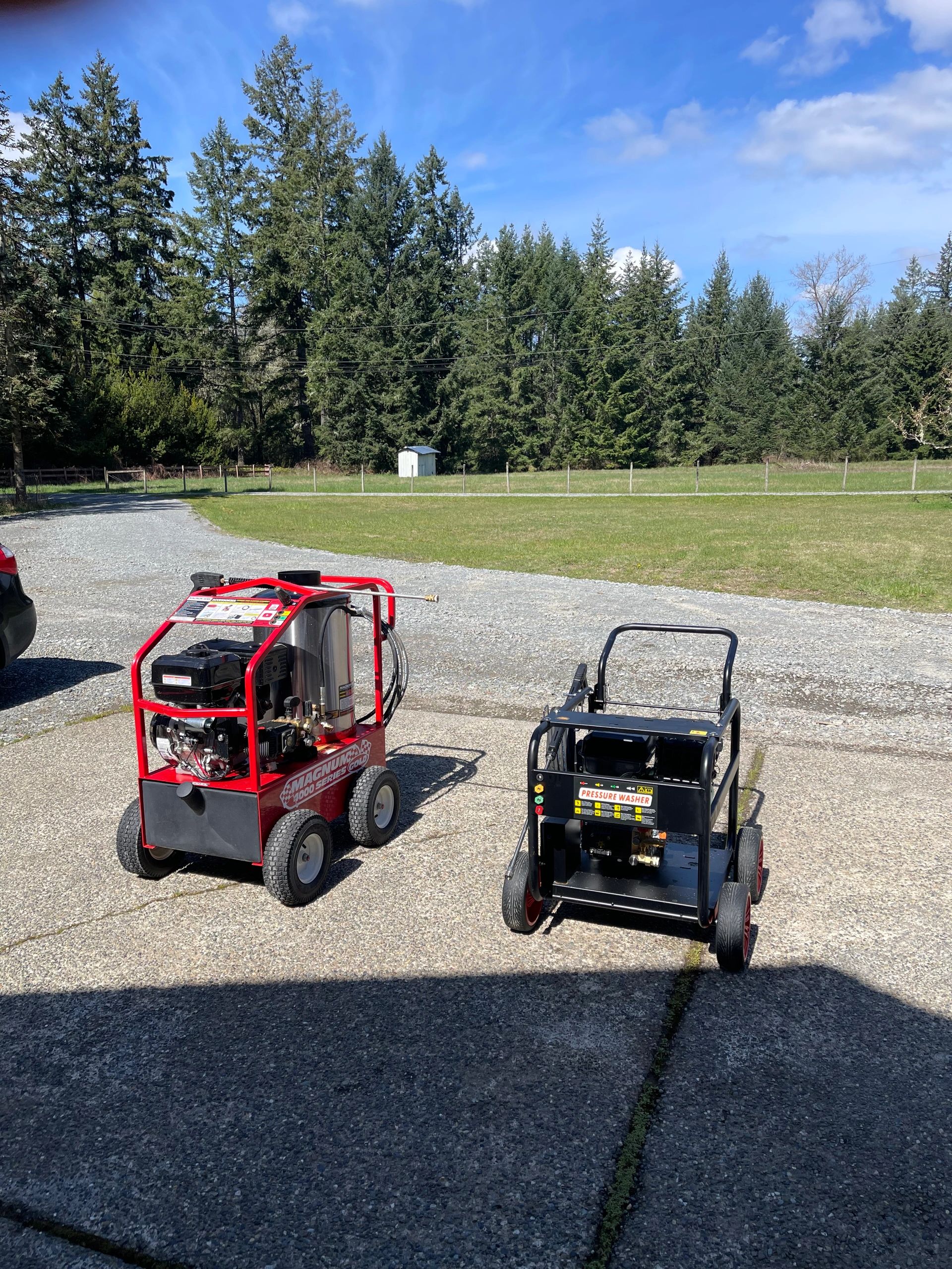 Two pressure washers on a sunny driveway with trees in the background.