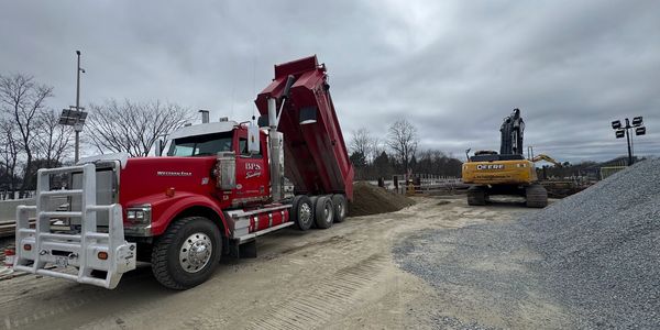 Red dump truck unloading gravel at construction site with excavator nearby.
