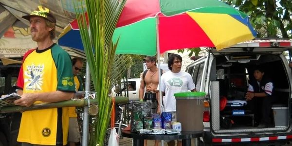 A vendor in a colorful shirt stands under a large umbrella